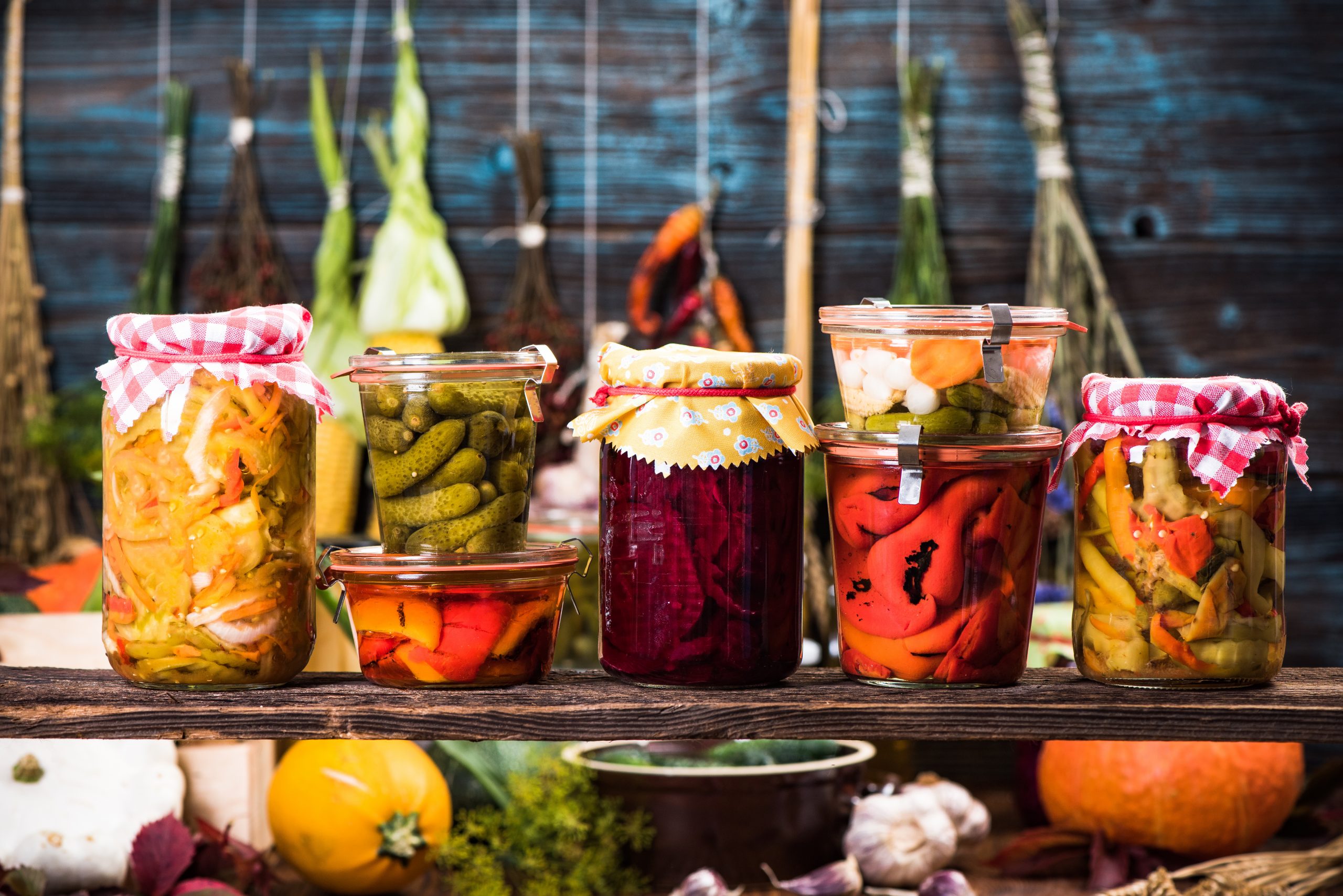 Various homemade pickles in glass jars on a wooden table, surrounded by fresh vegetables and garlic.