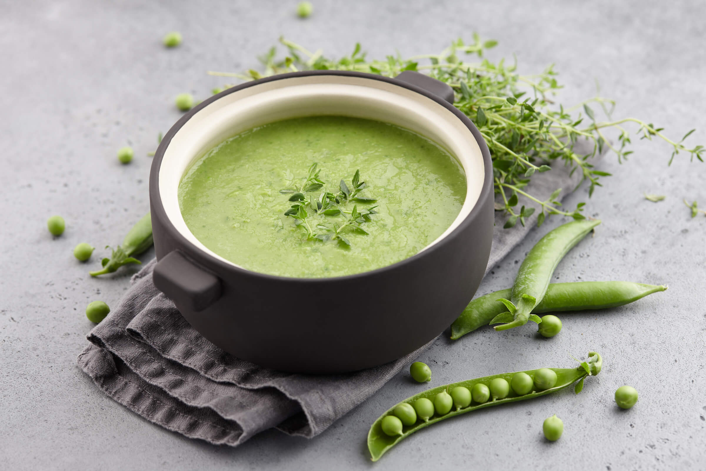 A bowl of fresh green pea soup garnished with herbs, surrounded by pea pods and sprigs of thyme on a gray background.